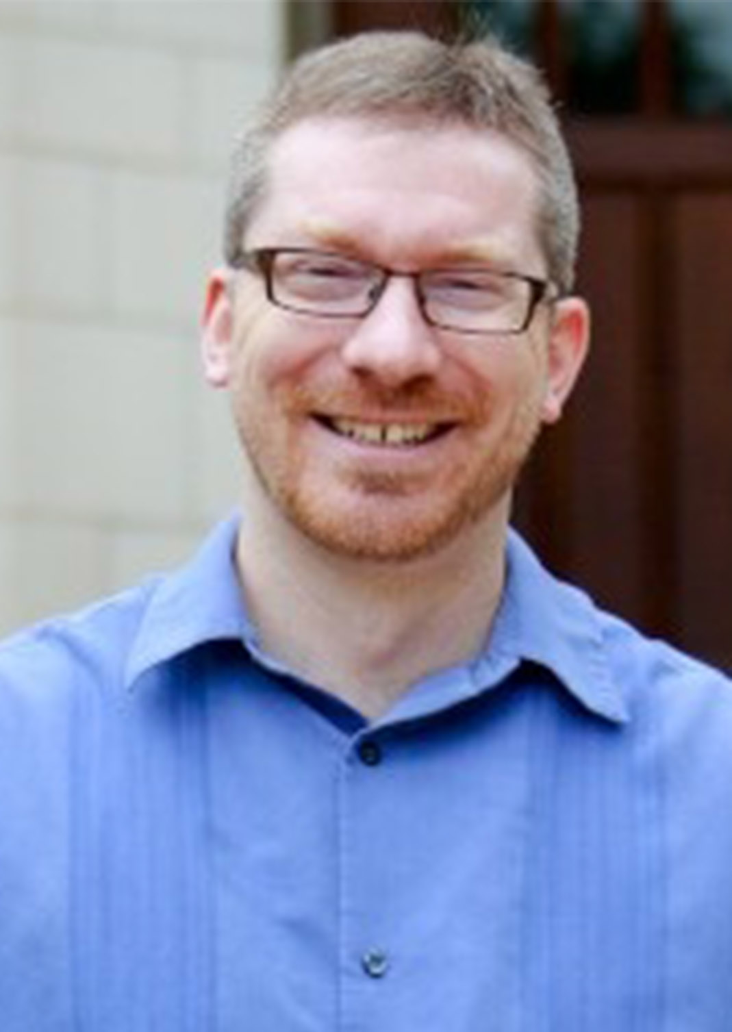J.P. Masly has short reddish hair, a goatee, and glasses and is smiling. He is outside and is wearing a blue, collared shirt. In the background is a gray, brick wall and a dark, wooden door.